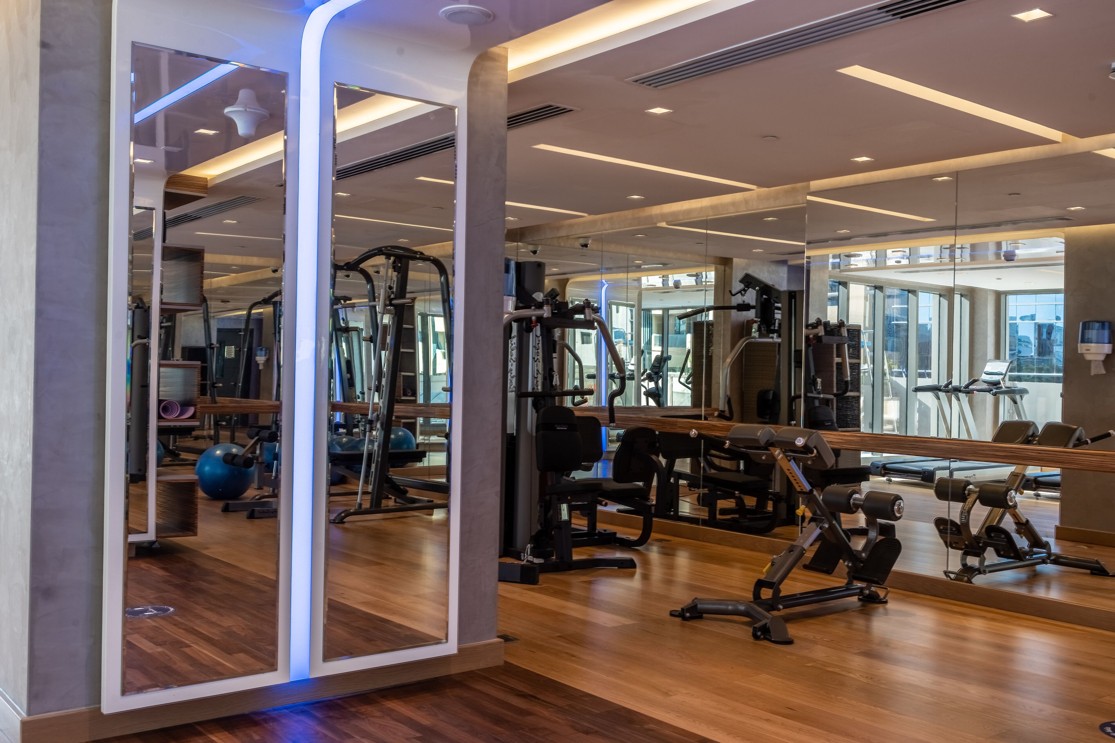 A modern gym with wooden flooring, various exercise equipment, and large mirrors.
