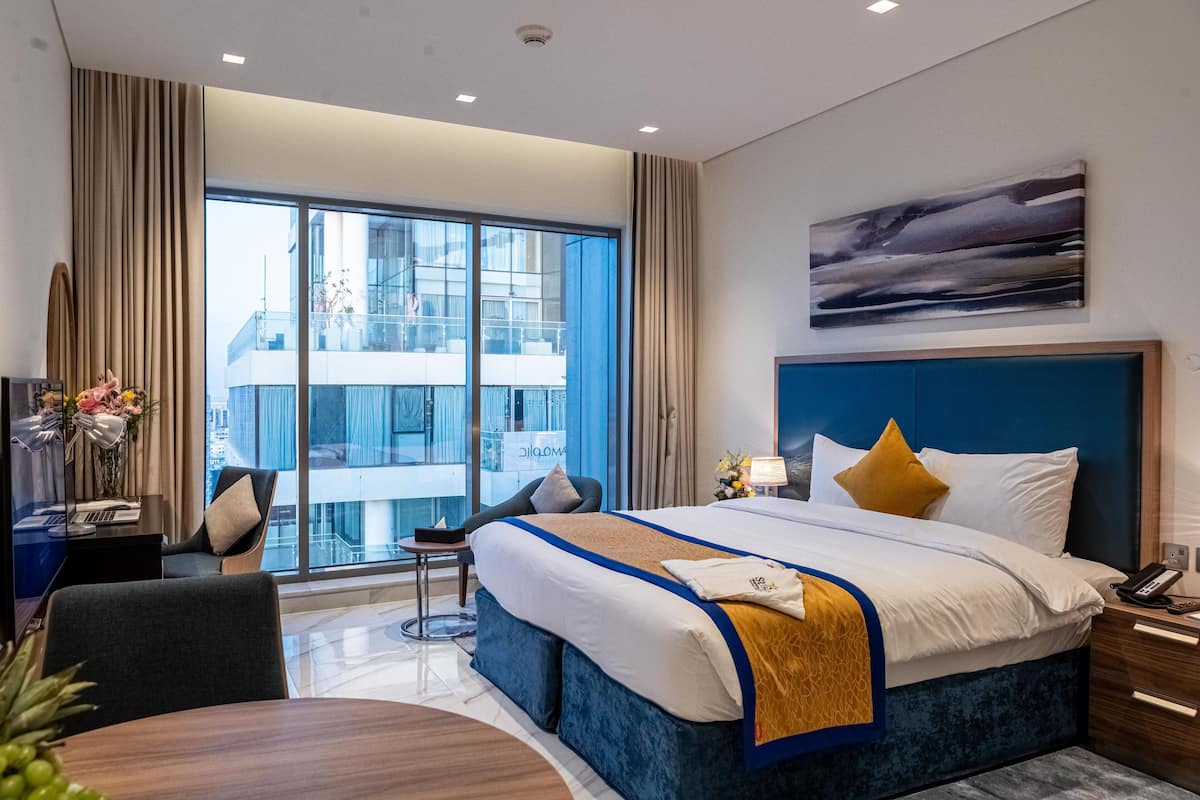 Une chambre d’hôtel moderne avec un grand lit, un bureau équipé d’un ordinateur, une chaise et une vue sur un immeuble grâce à de grandes fenêtres.