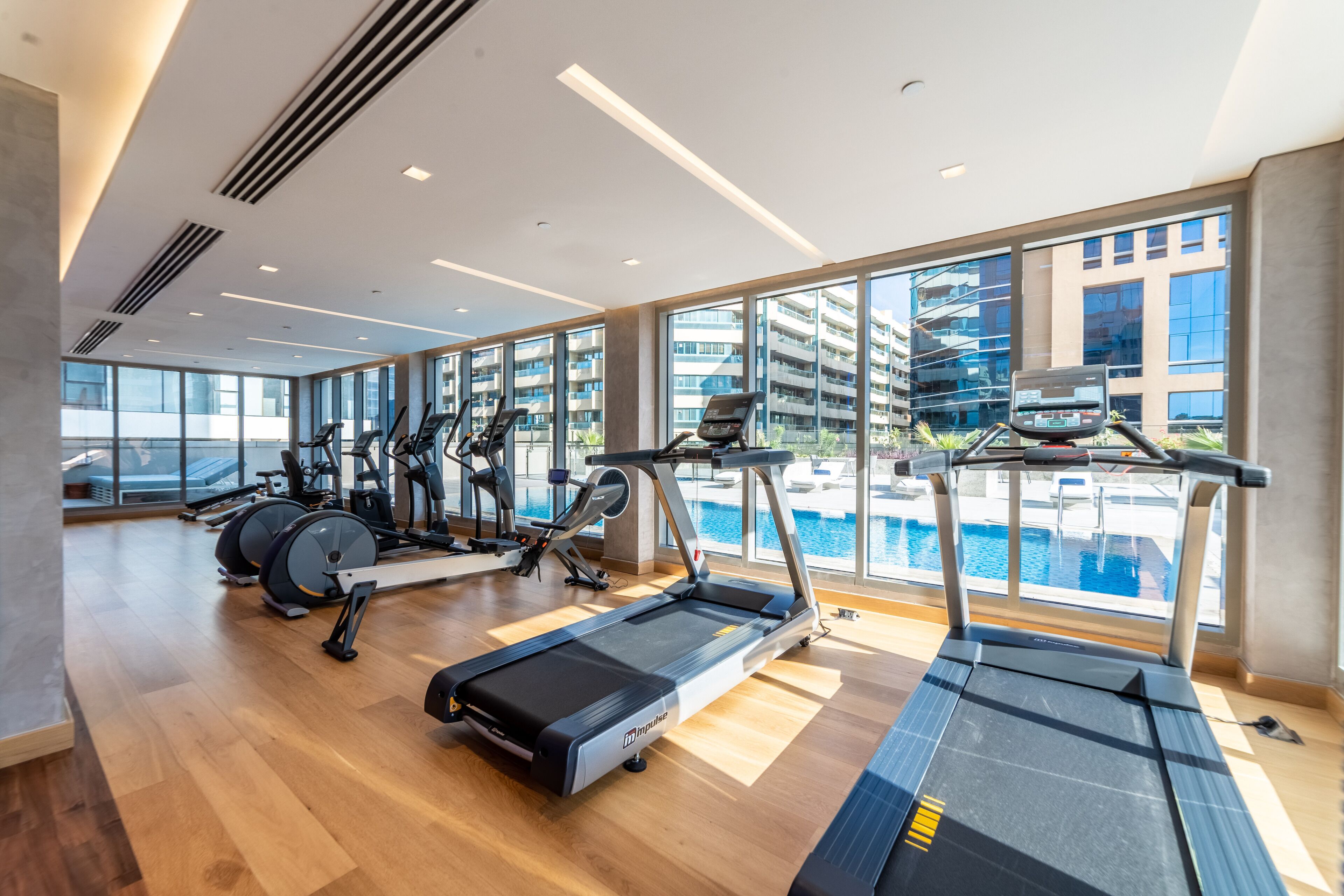 A modern gym with treadmills, elliptical machines, and rowing machines, large windows, and a pool view.