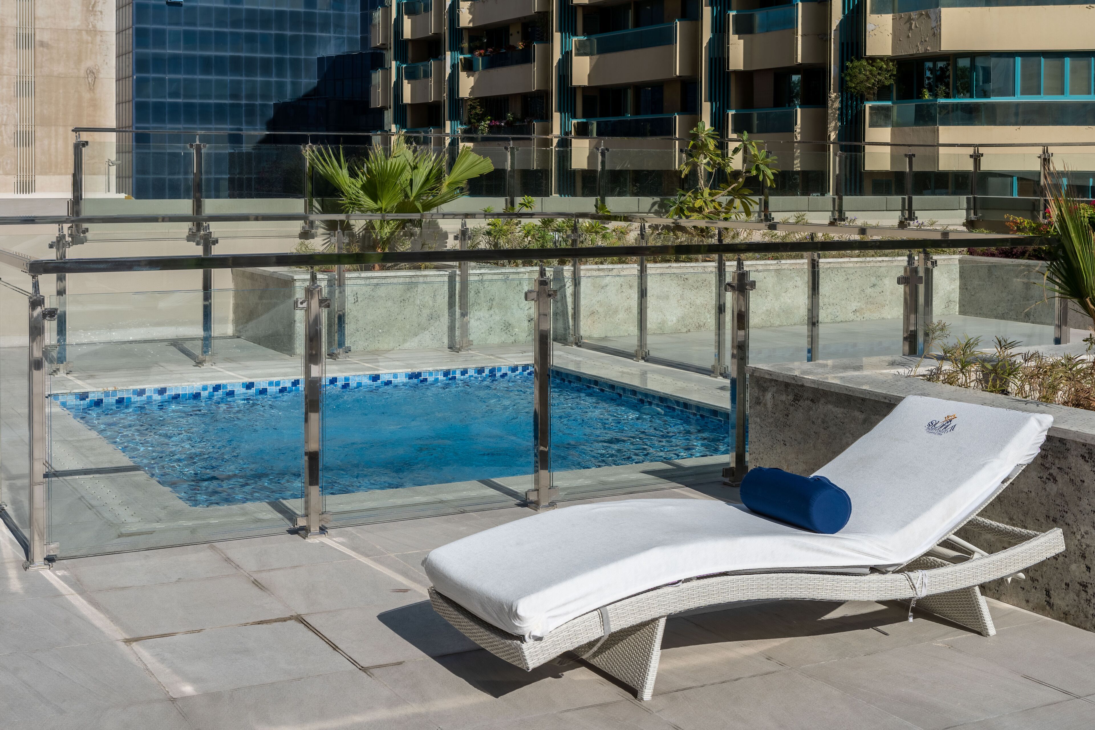 Zona de piscina en la azotea con una tumbona, barandillas de cristal y vistas a un edificio moderno.