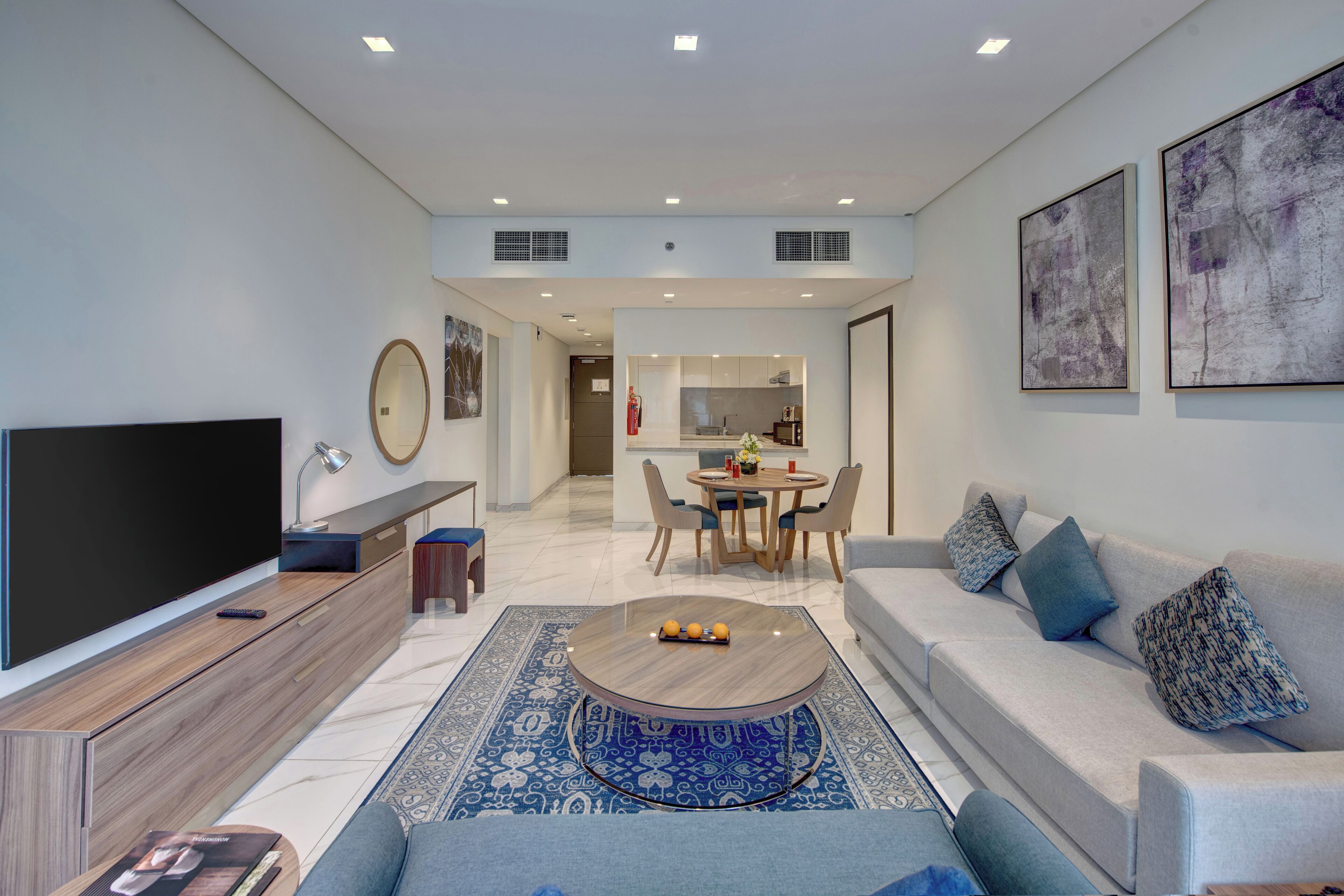 A modern living room with a flat-screen TV, a coffee table, and a sofa. There is a dining area with a table and chairs in the background.