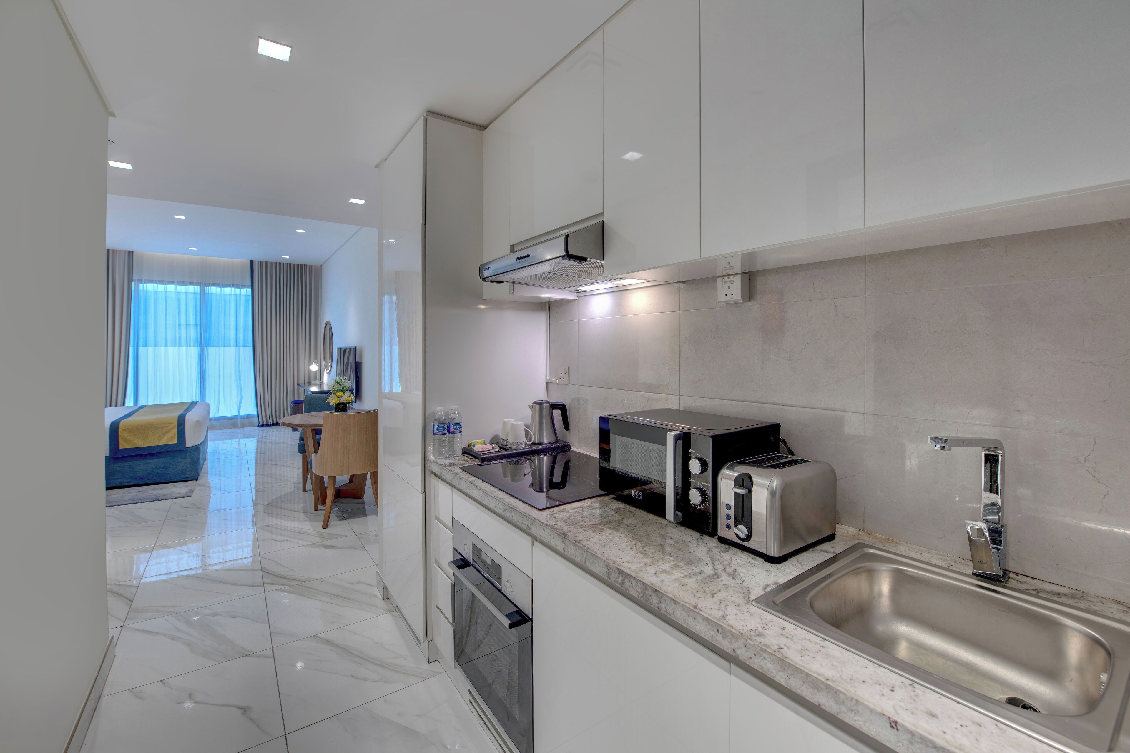 Una cocina moderna con un lavamanos de acero inoxidable, un microondas, un tostador y una cafetera. Se ve un área de comedor con una mesa y sillas al fondo.