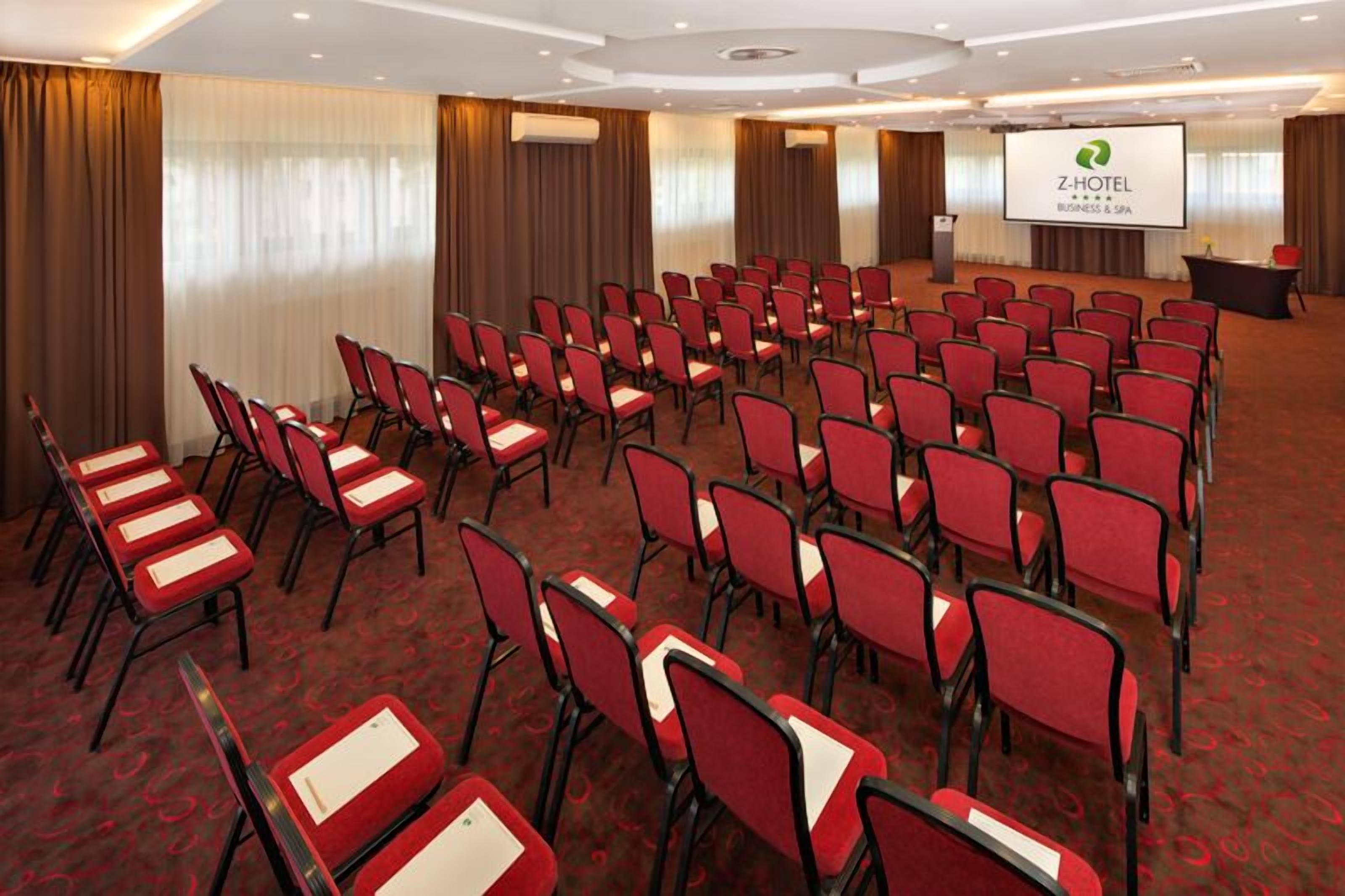 A conference room with rows of red chairs, a podium, and a screen displaying the Z-Hotel logo.