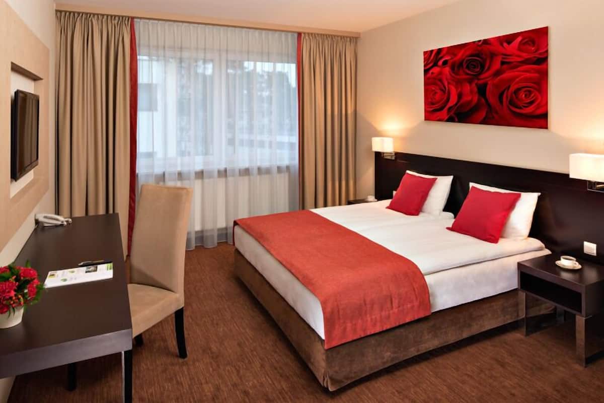 A hotel room with a bed, a desk, a chair, a nightstand, and a painting of red roses.