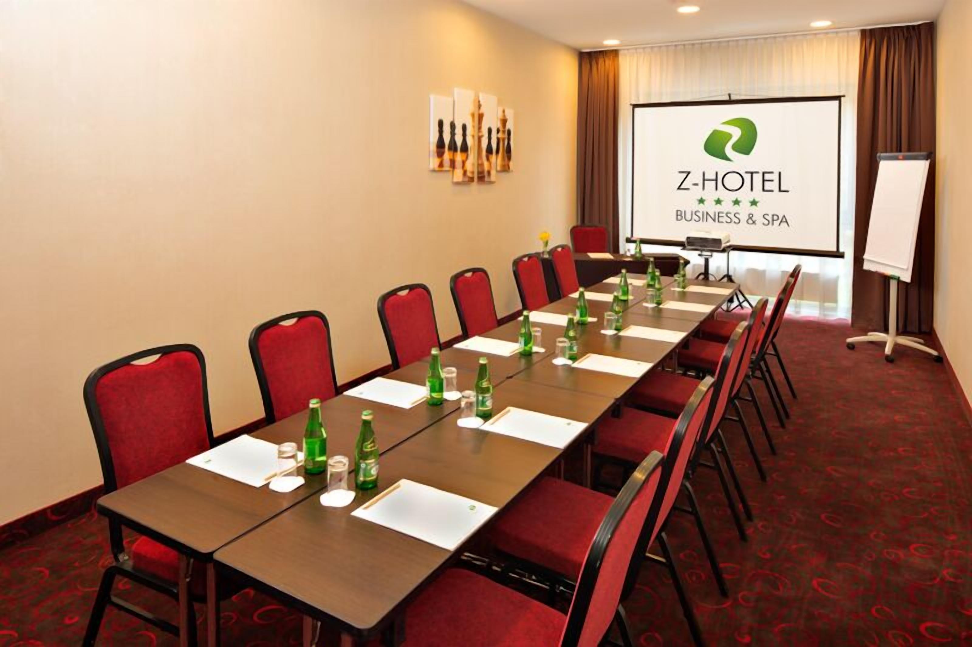 Ein Konferenzraum mit langem Tisch, roten Stühlen und einer Projektionsleinwand, auf der das Logo des Z-Hotel zu sehen ist.