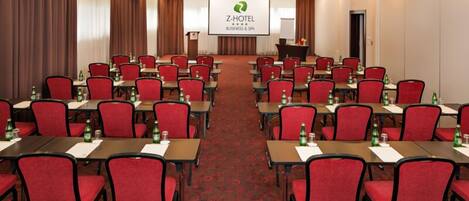 Un salón de conferencias con filas de sillas rojas y mesas, un atril y una pantalla mostrando el logotipo de Z-HOTEL.