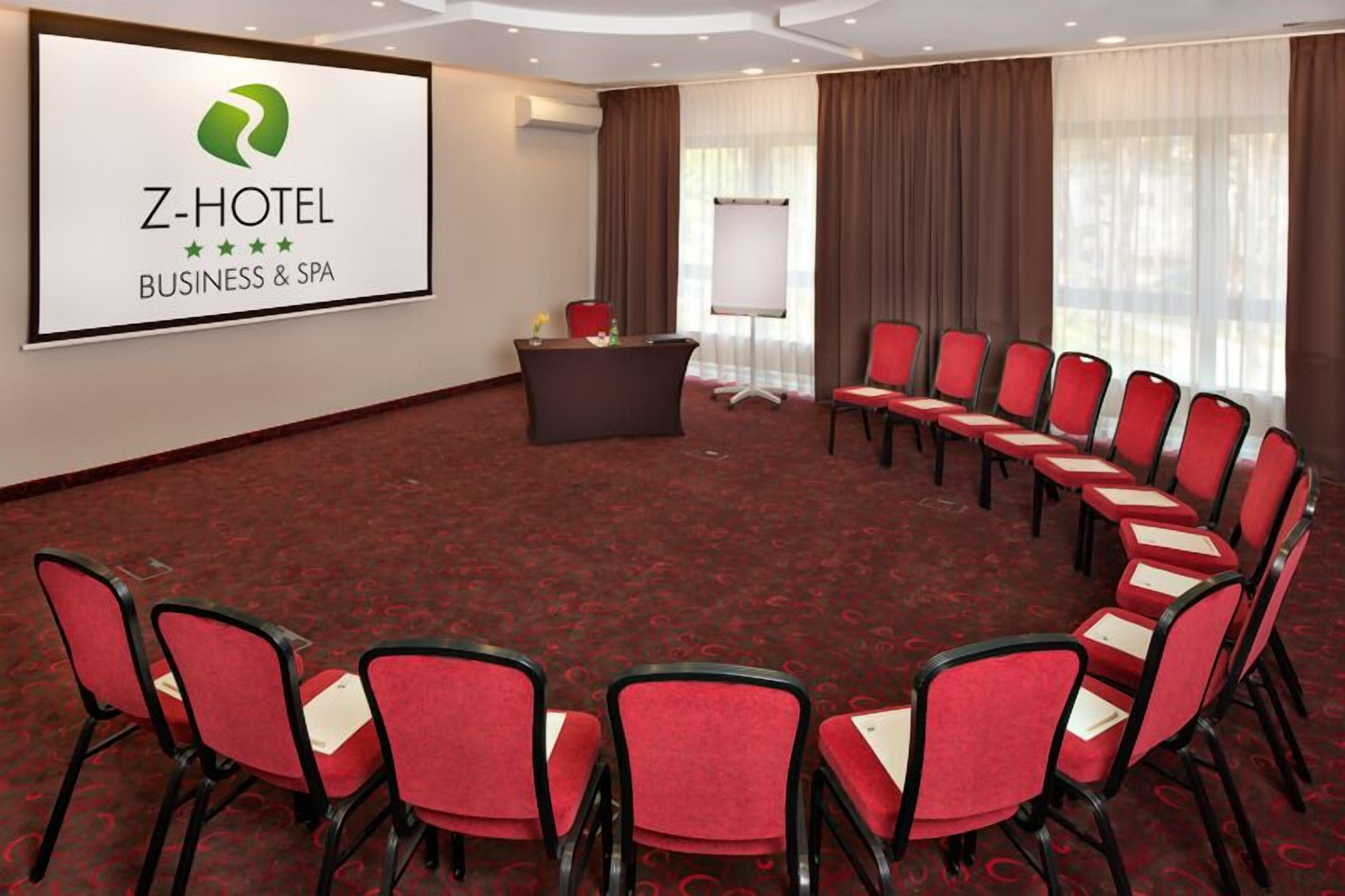 Een congreszaal van Z-Hotel met een groot scherm waarop de naam van het hotel en de diensten staan, een ronde tafel en rode stoelen eromheen.