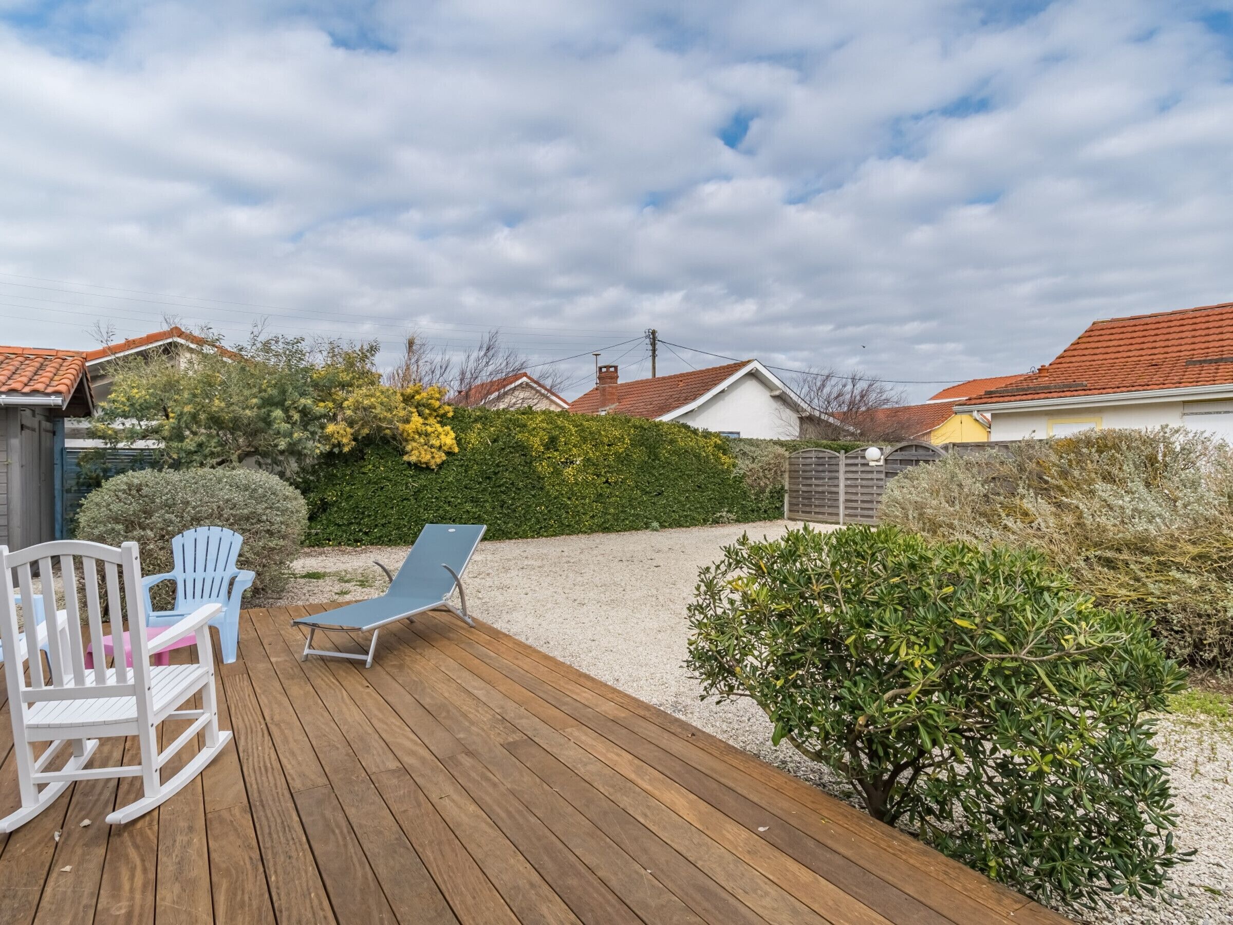 A wooden deck with two blue chairs and a rocking chair, surrounded by greenery and houses with red roofs.