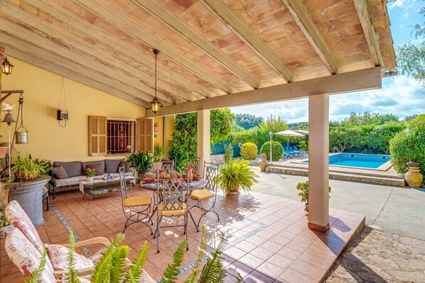 A covered patio area with a sofa, chairs, and a table, surrounded by potted plants and a view of a swimming pool.