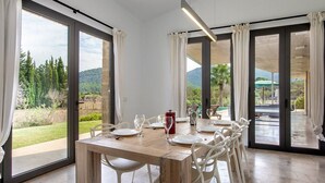 Un coin repas avec une table dressée pour six personnes, de grandes portes-fenêtres coulissantes et une vue sur un jardin et des montagnes.