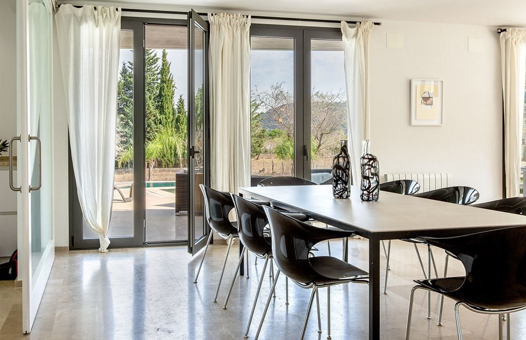 Un coin repas avec une table et des chaises, une vue sur les arbres et les montagnes à travers des portes vitrées, et un tableau encadré au mur.