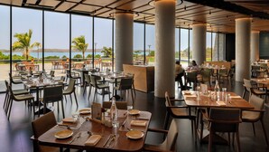 Un restaurante espacioso con techos altos, amplias ventanas y vistas a la playa. Las mesas están preparadas con platos, cubiertos y vasos.