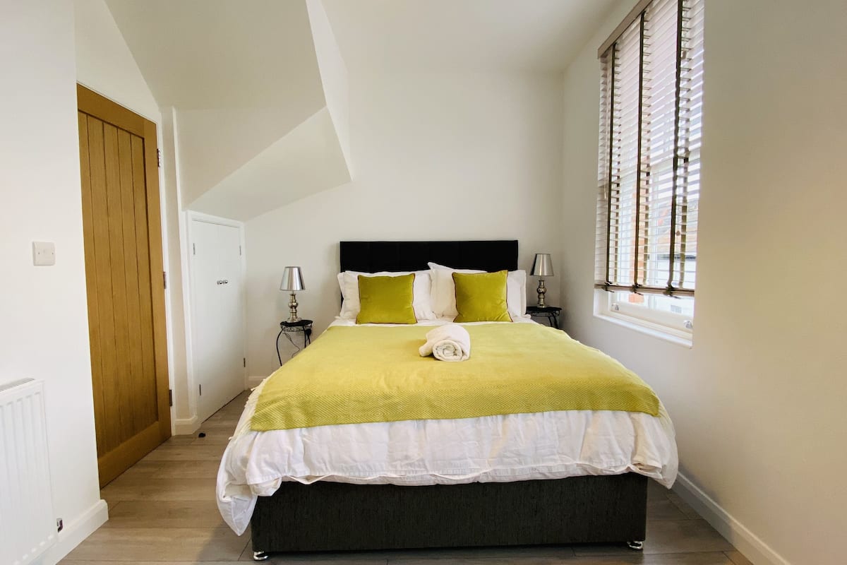 A bedroom with a bed, a window with blinds, a door, and a nightstand with a lamp.