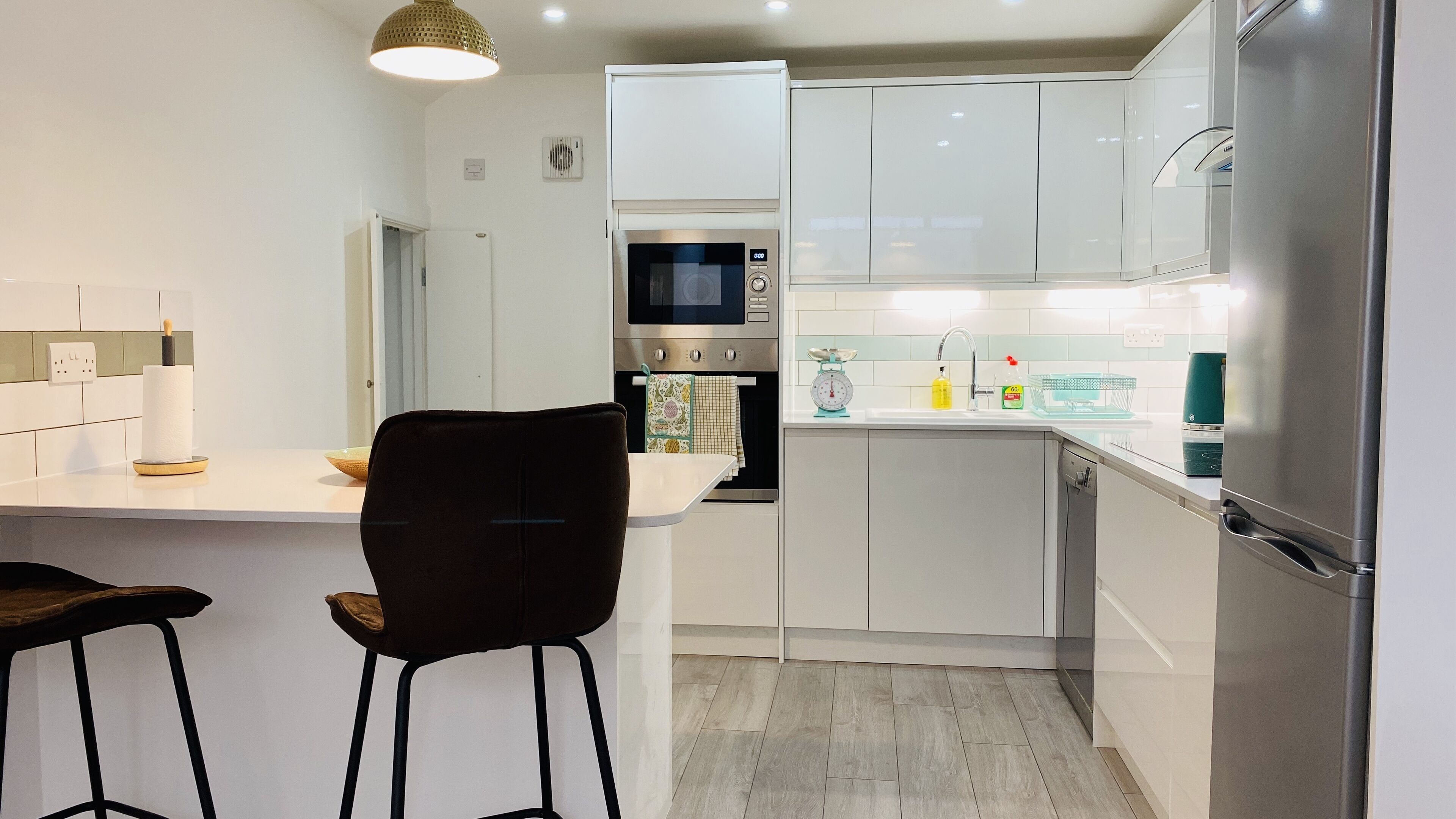 A modern kitchen with white cabinets, a built-in oven, a microwave, and a stainless steel refrigerator. There are two bar stools and a hanging towel.