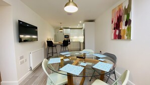 A modern dining area with a glass table surrounded by chairs, a kitchen in the background, and a colorful abstract painting on the wall.