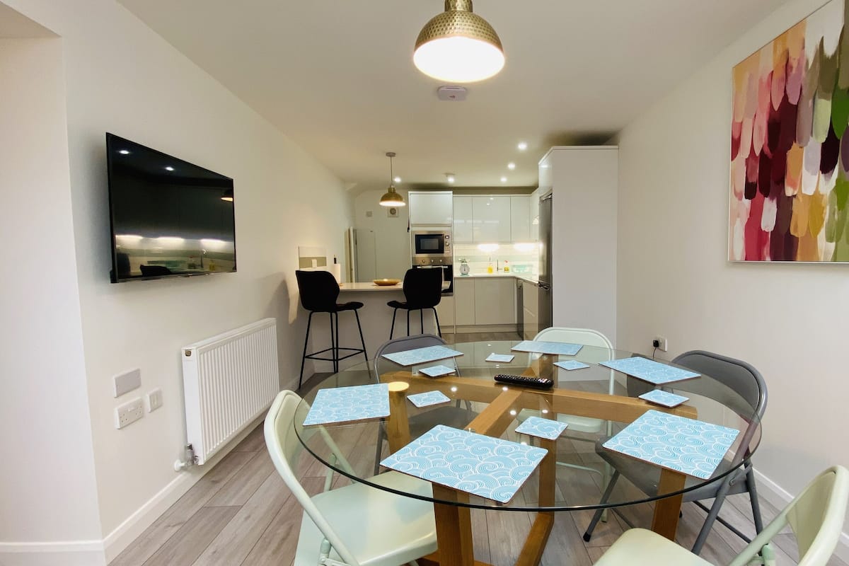 A modern dining area with a glass table surrounded by chairs, a kitchen in the background, and a colorful abstract painting on the wall.