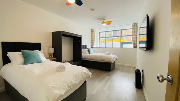 A hotel room with two beds, a wardrobe, a ceiling fan, a TV, and a window with a view of buildings.