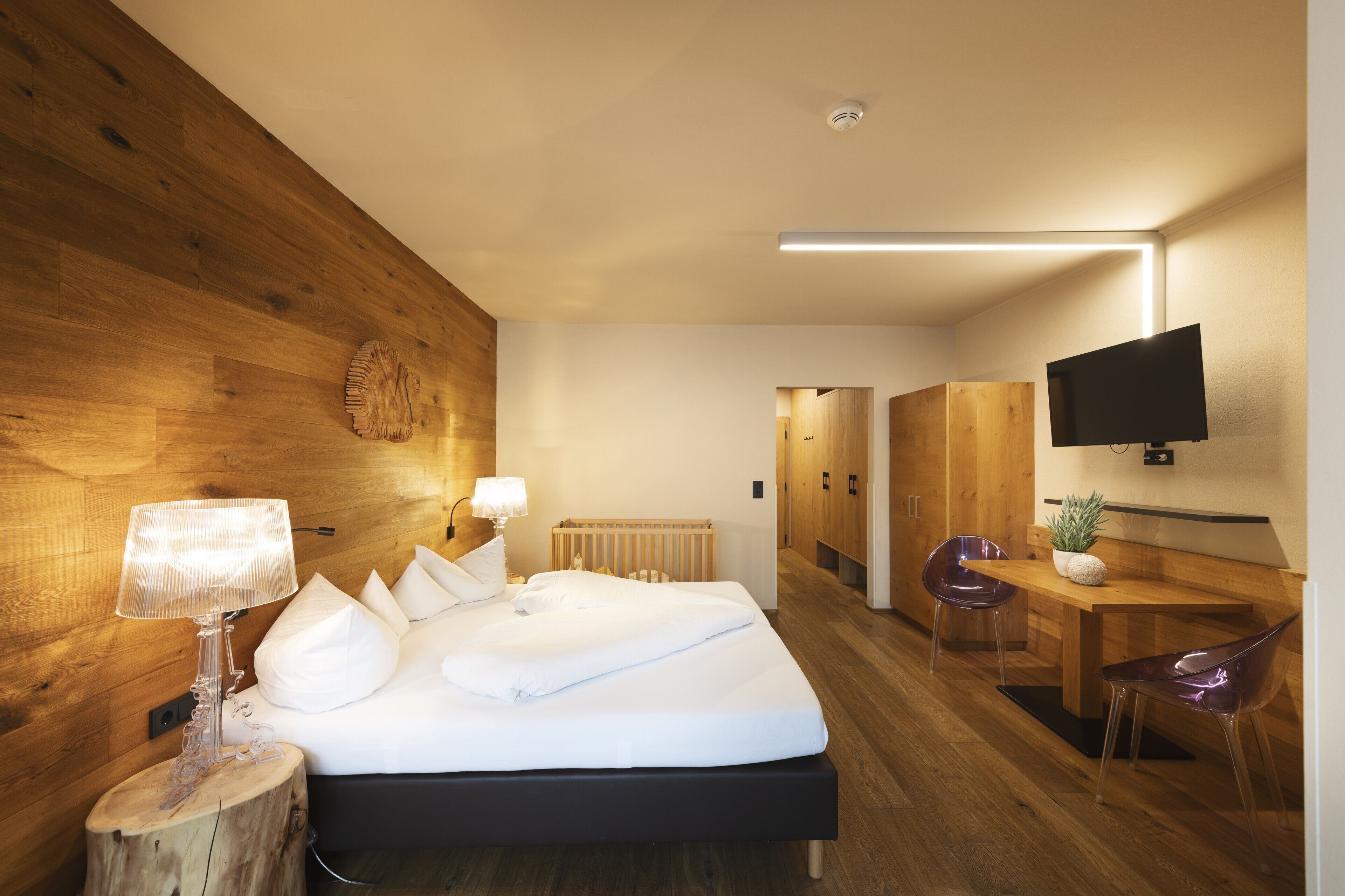 Una habitación de hotel moderna con una pared de madera, una cama con ropa blanca, un escritorio con silla y un televisor montado en la pared.