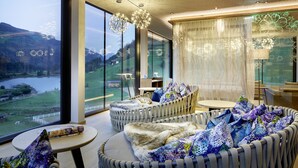 A modern interior with wicker furniture, large windows, and a view of a lake and mountains. - Almhof Family Resort & SPA (Gerlos)