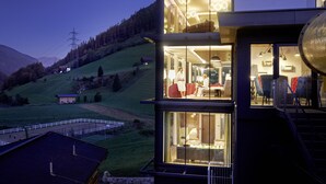 A modern two-story building with large glass windows, a dining area, and a view of a hilly landscape. - Almhof Family Resort & SPA (Gerlos)