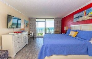 Ein Hotelzimmer mit Bett, Fernseher, Schreibtisch, Stuhl, einem Balkon mit Meerblick und einer roten Wand.