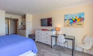 A hotel room with a bed, a desk, a TV, a painting, and a kitchenette.