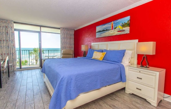A hotel room with a bed, a nightstand, a chair, a balcony with a view of the ocean, and a painting on the wall.