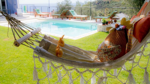 A hammock with a plush toy and a bottle, overlooking a pool with lounge chairs and umbrellas, set against a scenic hillside backdrop. - Il Sole di Poggi (Imperia)