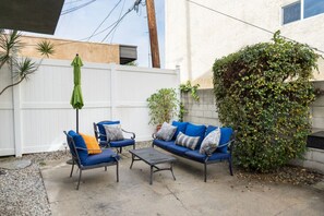 Une terrasse avec deux chaises bleues et une petite table, une clôture blanche et une grande plante à feuilles abondantes.