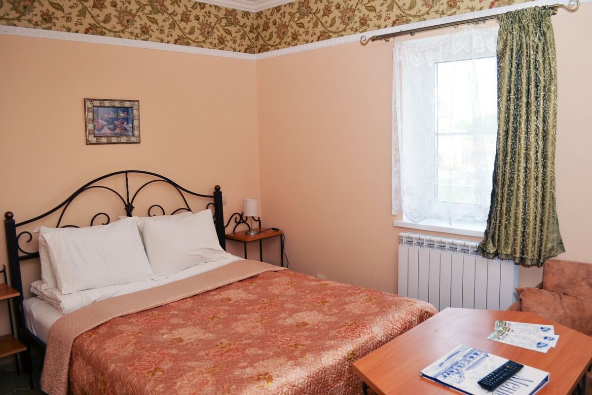 Un dormitorio con cama, mesita de noche, lámpara, ventana con cortinas, una silla y una mesa con papeles y un control remoto.