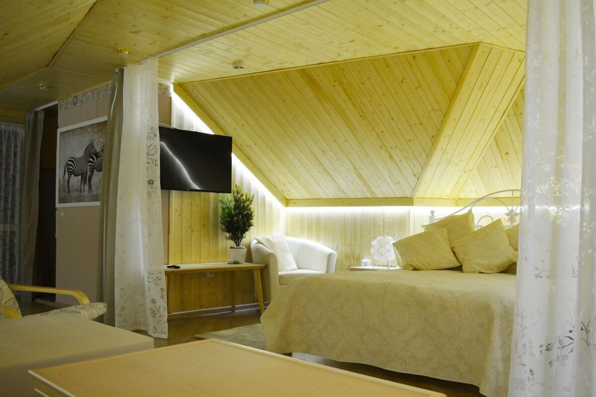 Una habitación acogedora y bien iluminada, con techo de madera, una cama con almohadas amarillas, una mesita pequeña y un televisor montado.