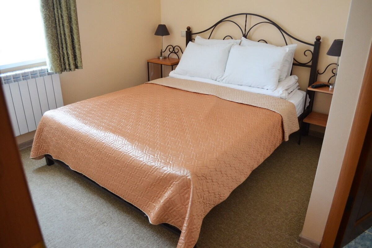 Una cama bien tendida con un edredón color beige, dos mesitas de noche con lámparas y una ventana con cortinas.