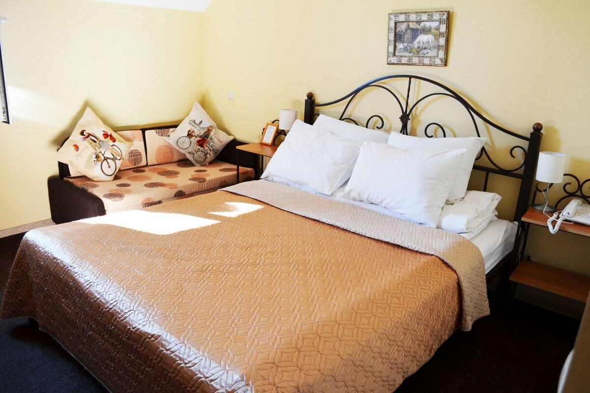 Una cama doble con colcha marrón, dos mesitas de noche con lámparas, un cuadro en la pared y una silla con cojines estampados.