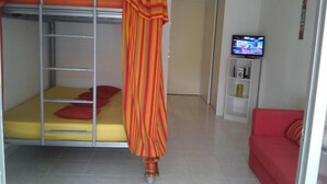 A dormitory-style room with a bunk bed, a red sofa, a television, and a small shelf with items. - Sun, sea and easy living in this studio close to everything (SAINT FRANCOIS)