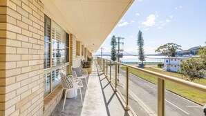 A balcony with a view of a coastal road, a building with a brick facade, and a clear sky. - Shoal Court 4, 7 Lillian St - water views , linen, bed making (Shoal Bay)