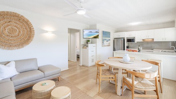 A modern kitchen with white cabinets, a dining area with a round table and chairs, a sofa, and a woven wall hanging. - Shoal Court 4, 7 Lillian St - water views , linen, bed making (Shoal Bay)