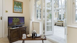 A room with a television on a wooden cabinet, a coffee maker, a plate of cookies, and a cup on a table. There is a door leading to an outdoor area with wicker furniture. - Chipping Campden Modern House, Garden, upstairs/downstairs bedrooms/bathrooms.  (Chipping Campden)