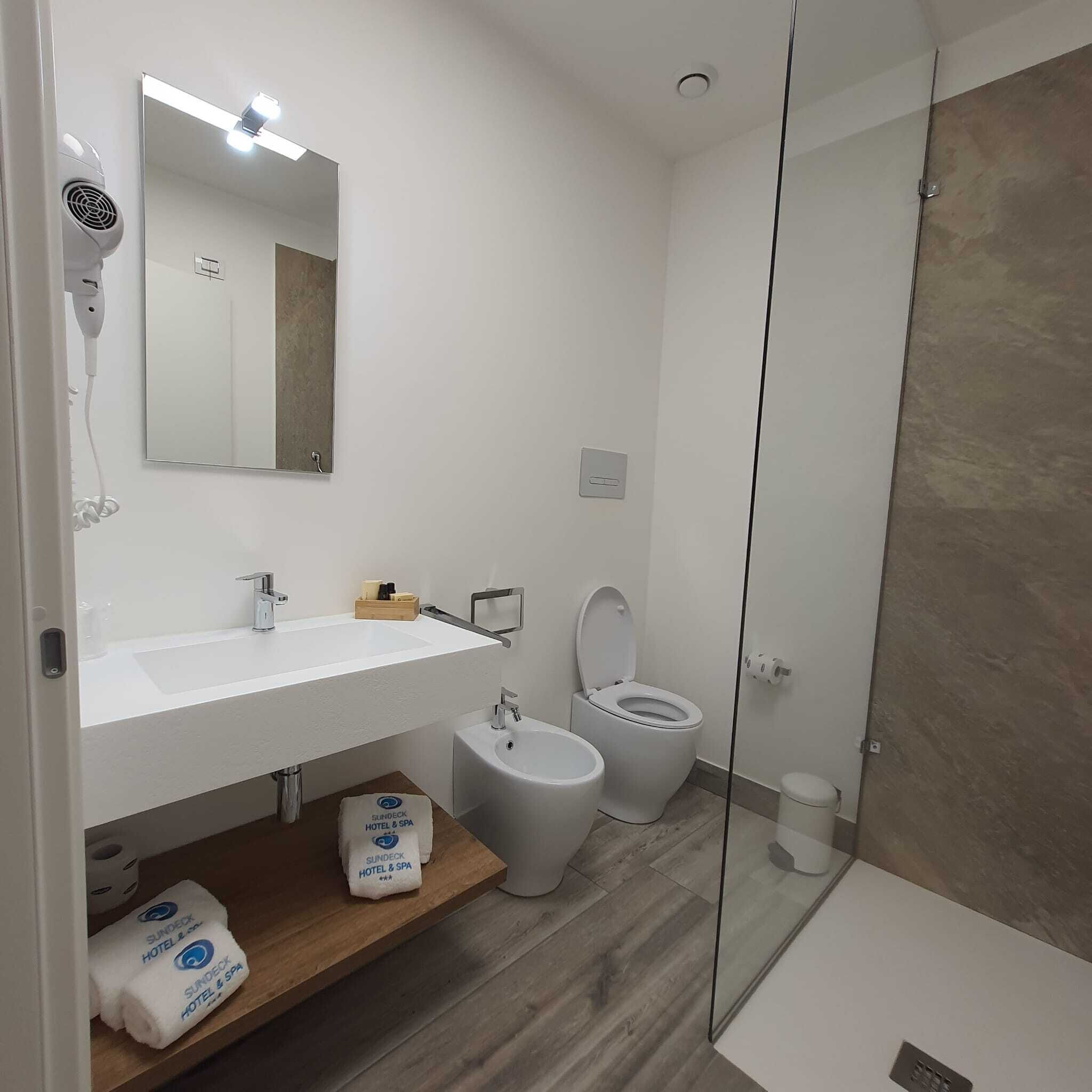Un bagno moderno con lavandino, specchio e servizi igienici bianchi. È presente un portasciugamani con asciugamani, una zona doccia e uno scaffale in legno con una borsa etichettata 'Sundeck Hotel & Spa'.