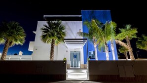 Un edificio moderno con fachada blanca y detalles azules, que presenta una entrada de vidrio y palmeras en primer plano.