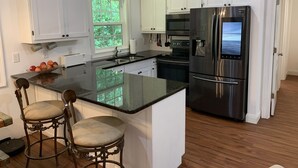 Une cuisine moderne avec des meubles blancs, des appareils en acier inoxydable, un grand îlot central doté de tabourets et une fenêtre donnant sur un espace verdoyant.
