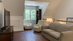Un salon comprenant un téléviseur, deux fauteuils blancs, un canapé beige, un plancher en bois, une fenêtre donnant sur un paysage verdoyant et une lampe.