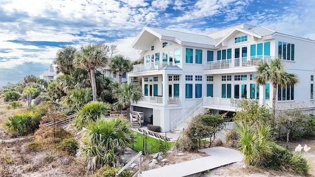 A multi-story beachfront house with a balcony, surrounded by palm trees and a rocky landscape. - 129 Dune Lane- Welcome to 5,000 square feet of oceanfront bliss! (Hilton Head Island)