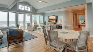 A spacious living room with a large window overlooking the ocean, a dining area with a round table, and a kitchen area with a television. - 129 Dune Lane- Welcome to 5,000 square feet of oceanfront bliss! (Hilton Head Island)