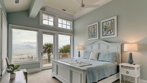 A bedroom with a large window overlooking a beach, a bed with a blue bedspread, and two wall-mounted lamps. - 129 Dune Lane- Welcome to 5,000 square feet of oceanfront bliss! (Hilton Head Island)
