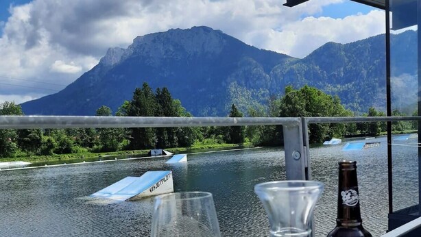 A table with a glass of white wine and a bottle of Zwetschgenbeer, a balcony overlooking a lake with boats and mountains in the background. - Ferienhaus Kaiserblick 125 qm für Max. 6 Personen und 3 Hunde (Kiefersfelden)