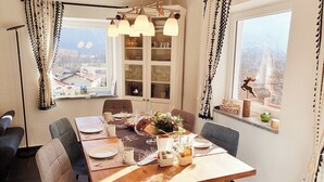 A dining area with a wooden table set for four, surrounded by chairs. A cabinet with glass doors is visible in the background, and the room has large windows with curtains. - Ferienhaus Kaiserblick 125 qm für Max. 6 Personen und 3 Hunde (Kiefersfelden)