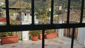Ein Balkon mit Fliesenboden und Terrakotta-Pflanzgefäßen, mit Blick auf eine hügelige Landschaft mit Gebäuden.