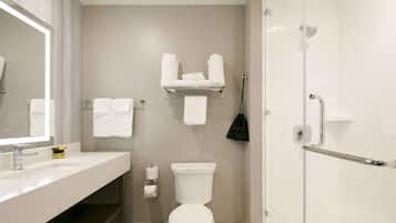 Une salle de bain moderne avec un lavabo, des toilettes et une douche, le tout de couleur blanche. On peut voir plusieurs serviettes blanches sur une étagère et un sac noir accroché au mur.
