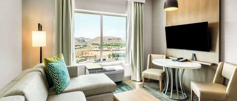 A modern hotel room with a large window, a flat-screen TV, a sofa, a coffee table, and a dining table.