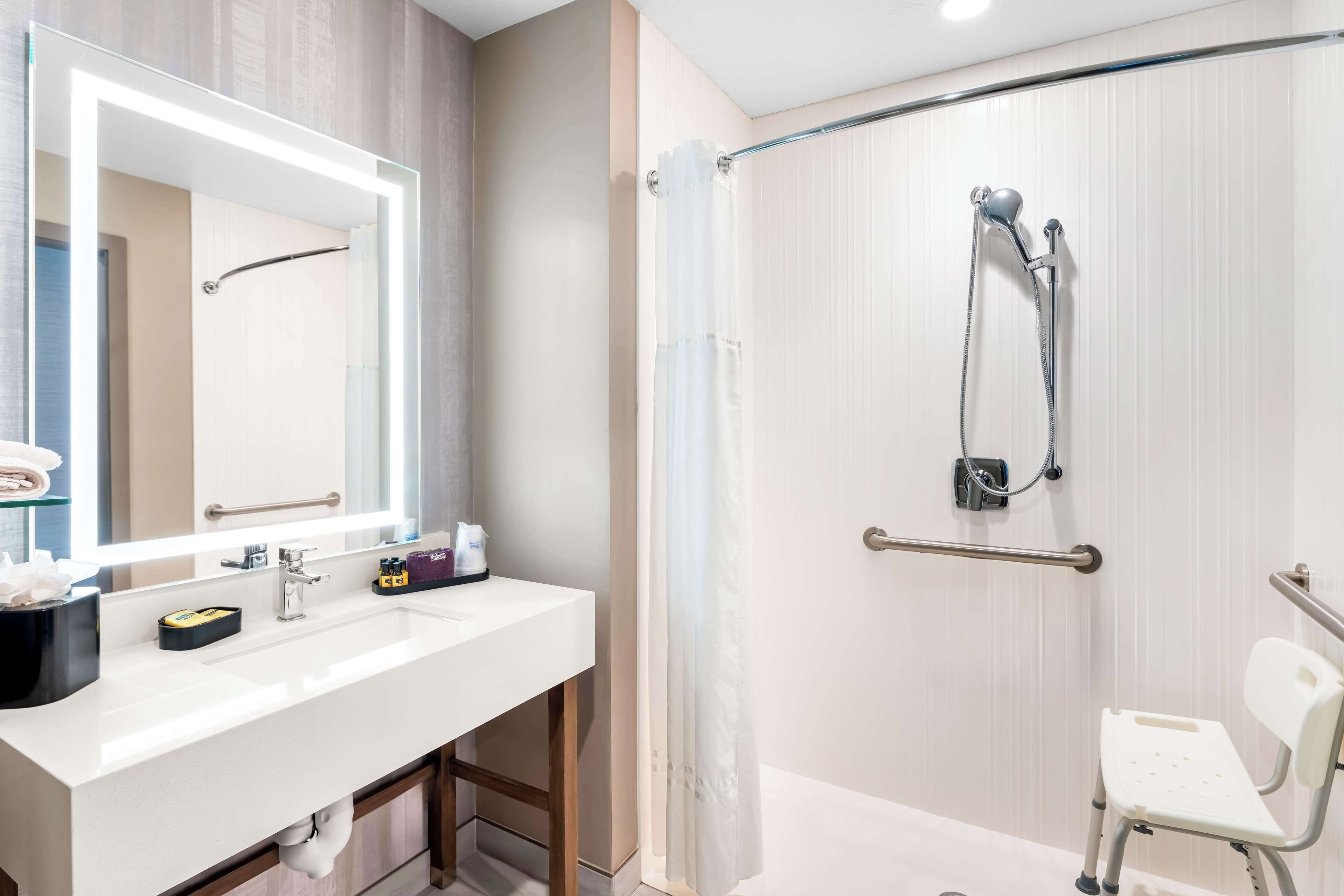 Une salle de bain moderne avec un lavabo blanc, un grand miroir, une douche avec une porte en verre et un banc accessible aux personnes en fauteuil roulant.